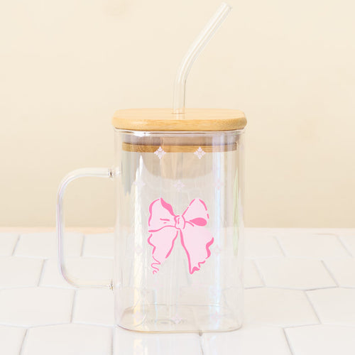 The Darling Effect’s Bow Square Glass Cup, a clear iridescent glass with handle, pink bow design, bamboo lid, and bent straw, sits on a white tiled surface against a light beige background.