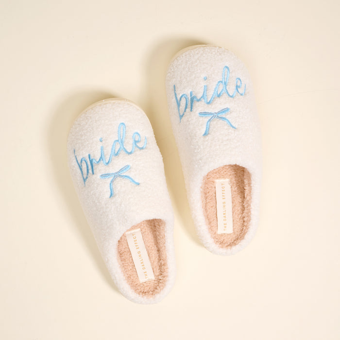 The Darling Effect Bride Fuzzy Slippers are cozy, white, and fuzzy with "bride" embroidered in blue and a small blue bow on each.