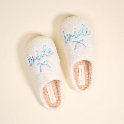 The Darling Effect Bride Fuzzy Slippers are cozy, white, and fuzzy with "bride" embroidered in blue and a small blue bow on each.