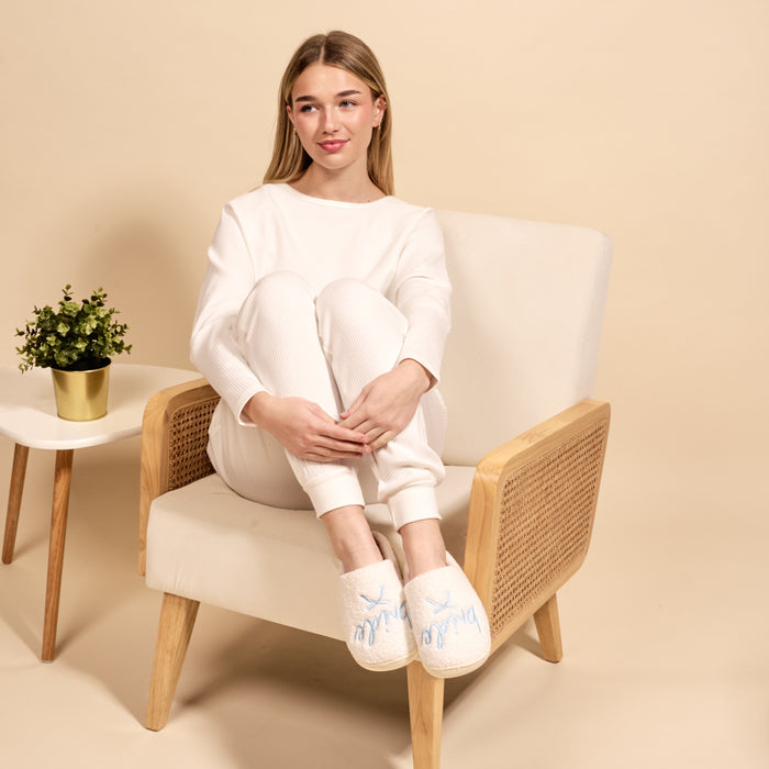 A woman in white loungewear sits on a beige chair, hugging her knees and wearing The Darling Effect’s Bride Fuzzy Slippers. A small potted plant sits on a white side table nearby, all against a neutral background.