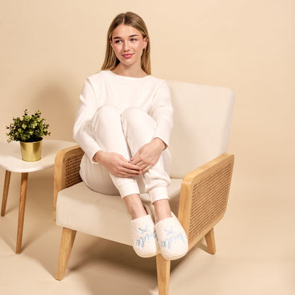 A woman in white loungewear sits on a beige chair, hugging her knees and wearing The Darling Effect’s Bride Fuzzy Slippers. A small potted plant sits on a white side table nearby, all against a neutral background.