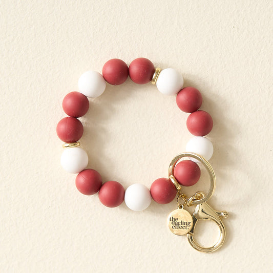 The Darling Effect’s Burgundy Gameday Hands-Free Keychain Wristlet features alternating burgundy and white round beads, a gold keyring, lobster clasp, and engraved gold charm—perfect as a game day wristlet or silicone bead keychain.