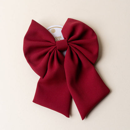 The Darling Effect's Burgundy Hair Bow—a large, deep red accessory with two long tails—comes attached to a white circular card and is displayed on a light beige background, making it a timeless piece for any occasion.