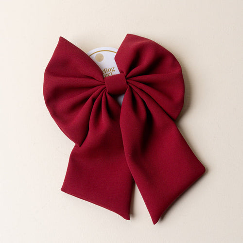 The Darling Effect's Burgundy Hair Bow—a large, deep red accessory with two long tails—comes attached to a white circular card and is displayed on a light beige background, making it a timeless piece for any occasion.