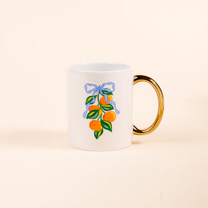 The Darling Effect’s Capri Citrus 11 oz Cozy Cute Ceramic Mug features a white ceramic body with a golden handle and showcases oranges and green leaves tied with a blue ribbon on the front, perfect for coffee or tea.