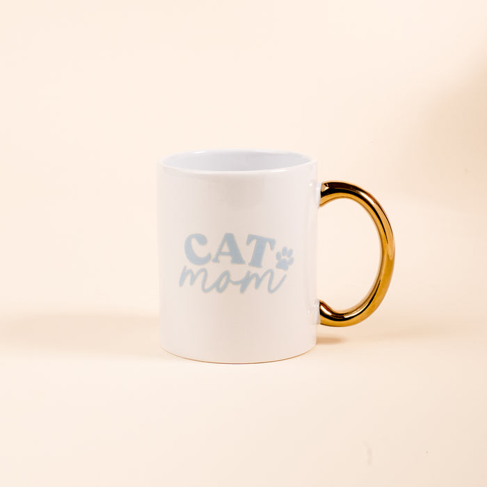 The Darling Effect’s Cat Mom 11 oz Cozy Cute Ceramic Mug features a white ceramic body, golden handle, and light blue "CAT mom" text with a paw print—ideal for coffee or tea. Shown on a plain light beige background.