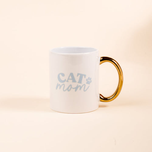The Darling Effect’s Cat Mom 11 oz Cozy Cute Ceramic Mug features a white ceramic body, golden handle, and light blue "CAT mom" text with a paw print—ideal for coffee or tea. Shown on a plain light beige background.