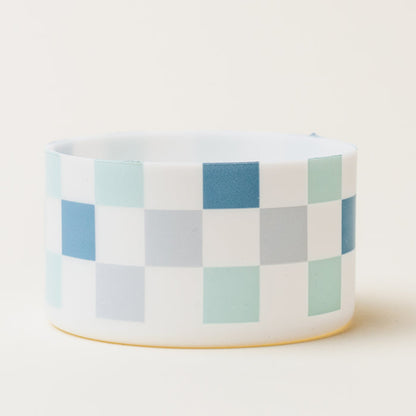 The Darling Effect’s Check Blue Tumbler Silicone Boot is a white cylindrical bowl with blue, green, and gray checkered squares, featuring a protective silicone boot.
