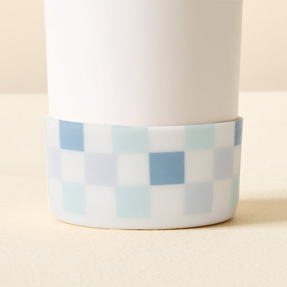 The Darling Effect Check Blue Tumbler Silicone Boot features a white cylindrical tumbler with a blue, teal, and white checkerboard silicone boot for added protection, shown on a light cream surface.