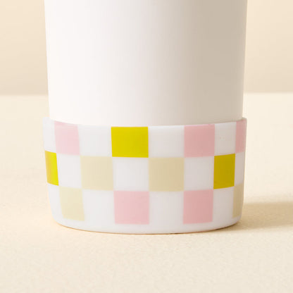 A close-up of The Darling Effect's Check Green Tumbler Silicone Boot, a white cylindrical tumbler accessory with a checkered silicone base in green, pink, yellow, beige, and white for added protection on a neutral surface.