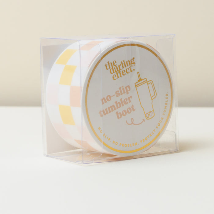 The Darling Effect’s Check Peach Tumbler Silicone Boot, featuring a round yellow and white check pattern, is displayed in a clear box with its label visible—stylishly protecting your tumbler.