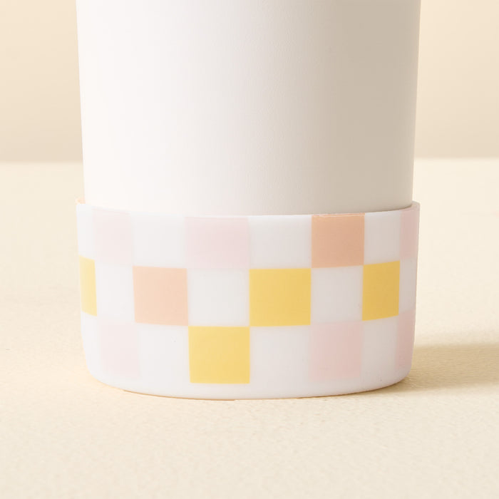 A close-up of The Darling Effect's Check Peach Tumbler Silicone Boot shows its pastel pink, yellow, and white checkered pattern. This accessory protects your tumbler while complementing light-colored surfaces and backgrounds.