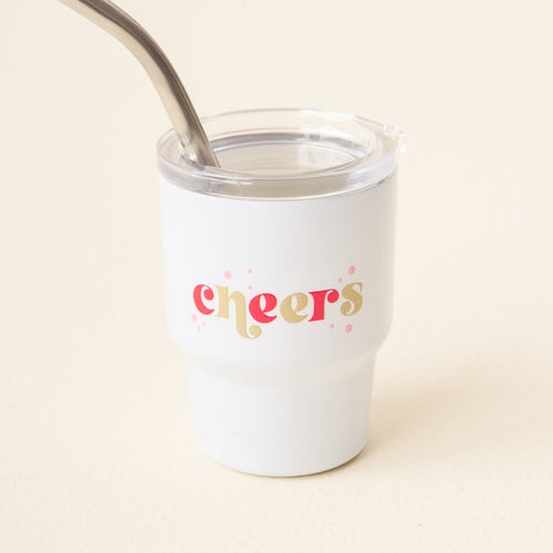 The Darling Effect Cheers 2 oz Tiny Tumbler features "cheers" in pink and gold script with pink confetti. Includes a metal straw extending from the lid.