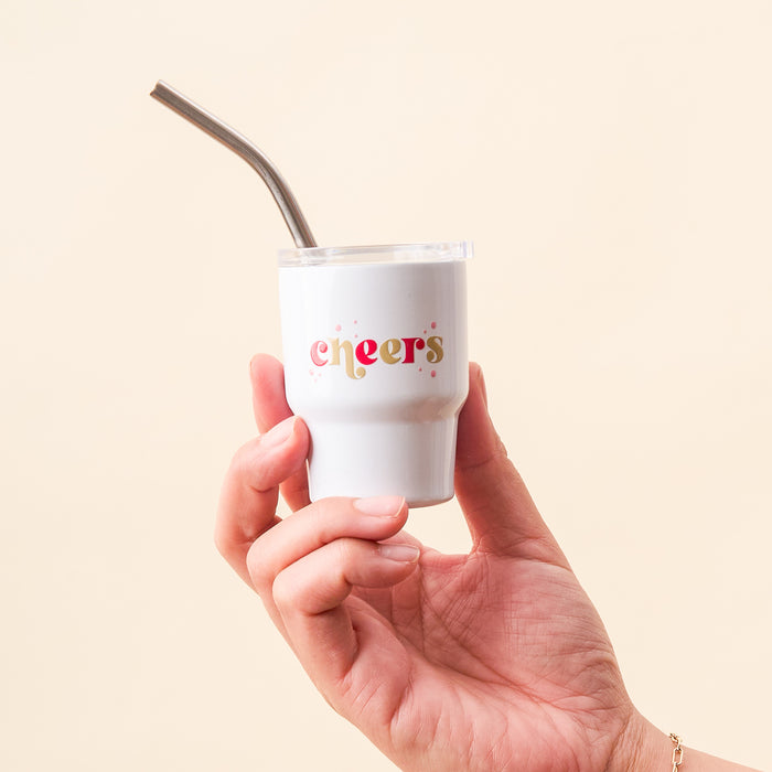A hand holds The Darling Effect's Cheers 2 oz Tiny Tumbler, a white insulated cup with a metal straw. The front features "cheers" in colorful letters, set against a light beige background.