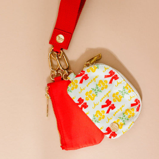 The Darling Effect’s Chi Omega Dynamic Duo Pouch Wristlet features a red wristlet with gold hardware, holding a red zip pouch and a white coin purse adorned with yellow-green flowers, red bows, and "hi" in yellow for stylish, hands-free use.