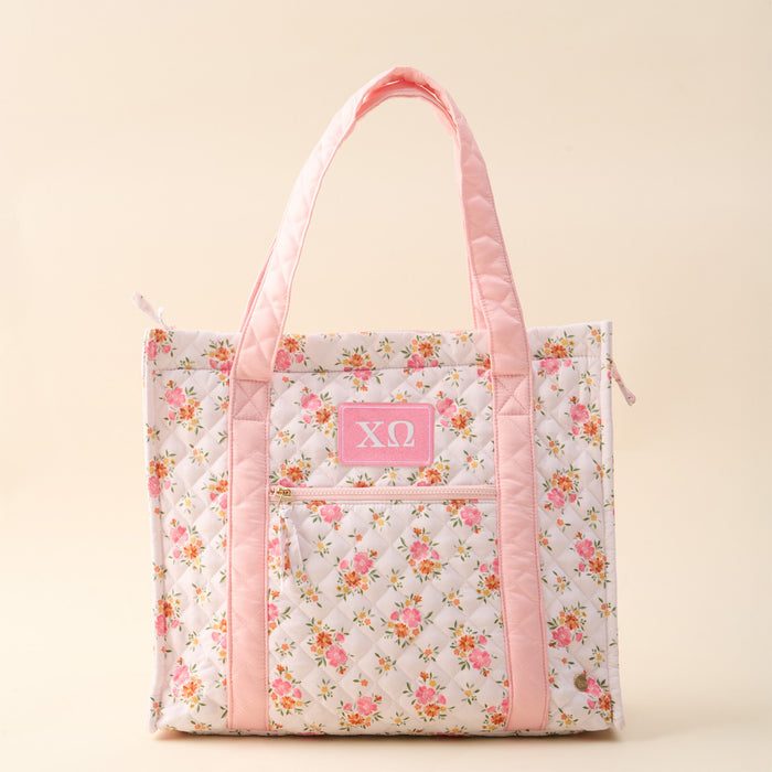 The Darling Effect’s Chi Omega The Everyday Quilted Tote Bag features pink handles, floral pattern, and a pink ΧΩ patch on the front pocket—an essential, stylish sorority tote for daily use.