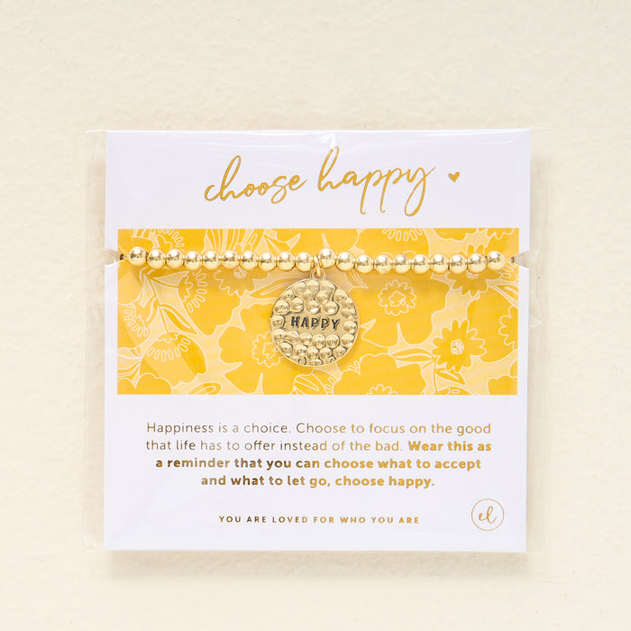 The Darling Effect's Choose Happy Beaded Charm Bracelet, featuring a gold beaded band and round charm with "HAPPY," is displayed on a yellow floral card—a thoughtful gift to inspire positivity and happiness each day.