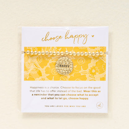 The Darling Effect's Choose Happy Beaded Charm Bracelet, featuring a gold beaded band and round charm with "HAPPY," is displayed on a yellow floral card—a thoughtful gift to inspire positivity and happiness each day.