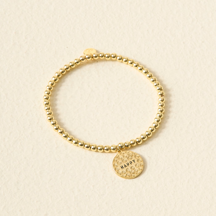 The Darling Effect’s Choose Happy Beaded Charm Bracelet features gold beads and a round, textured HAPPY charm—making it a meaningful gift, shown against a light beige background.