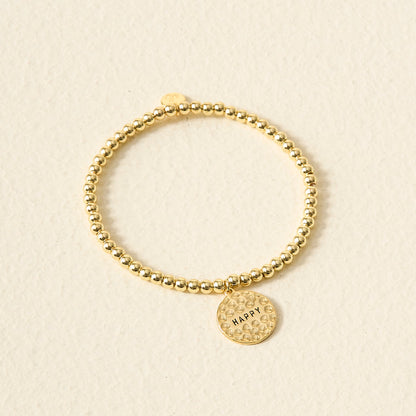 The Darling Effect’s Choose Happy Beaded Charm Bracelet features gold beads and a round, textured HAPPY charm—making it a meaningful gift, shown against a light beige background.