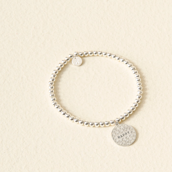 A silver Choose Happy Beaded Charm Bracelet from The Darling Effect rests on a light beige background. Two round charms—one plain, one engraved with "HAPPY" and dotted—make this a thoughtful, meaningful gift.