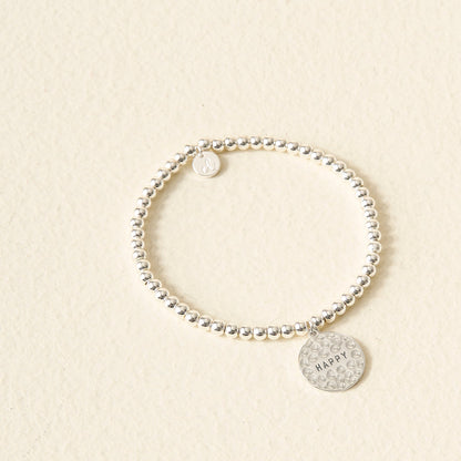 A silver Choose Happy Beaded Charm Bracelet from The Darling Effect rests on a light beige background. Two round charms—one plain, one engraved with "HAPPY" and dotted—make this a thoughtful, meaningful gift.