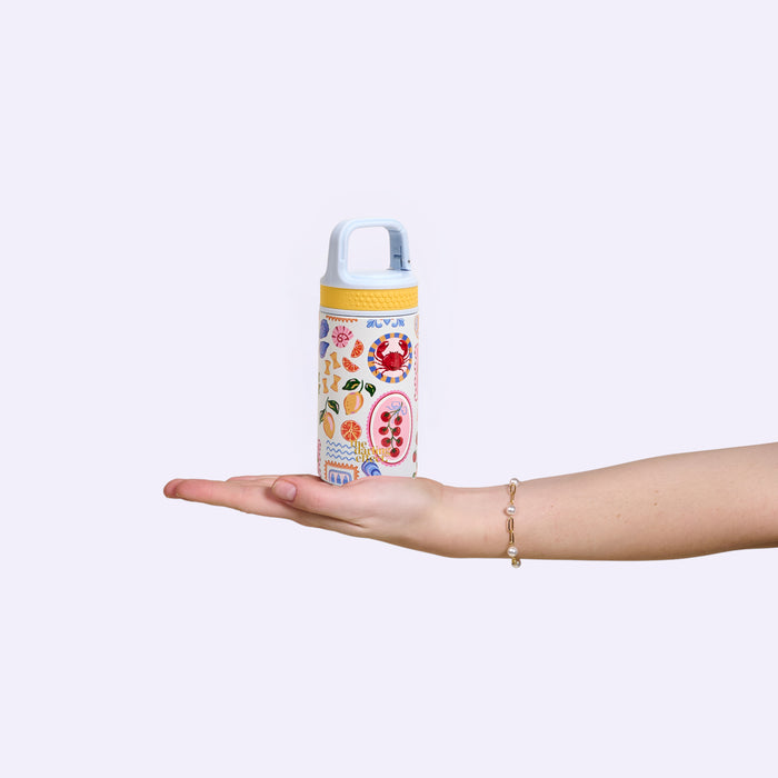 A hand holds The Darling Effect's Ciao Bella 6 oz Lil' Bitty Bottle, decorated with illustrated fruit and food stickers, against a light purple background—perfect for on-the-go hydration.