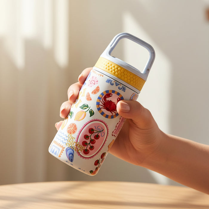 A hand holds The Darling Effect's Ciao Bella 6 oz Lil' Bitty Bottle, featuring a yellow lid, handle, and playful fruit and geometric designs. Sunlight casts gentle shadows on a light wooden table—perfect for on-the-go hydration.