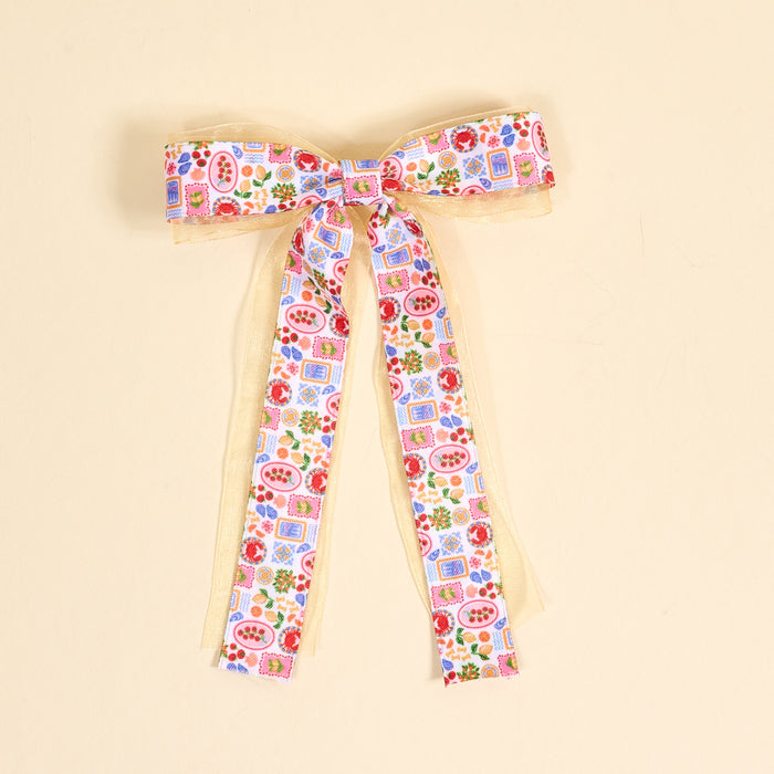 The Darling Effect's Ciao Bella Lovely Layered Hair Bow features long tails and a vibrant fruit, floral, and blue square pattern on white over gold sheer ribbon—an elegant accessory for standout hair looks. Photographed on a light beige surface.