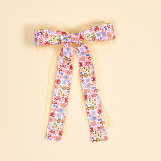 The Darling Effect's Ciao Bella Lovely Layered Hair Bow features long tails and a vibrant fruit, floral, and blue square pattern on white over gold sheer ribbon—an elegant accessory for standout hair looks. Photographed on a light beige surface.