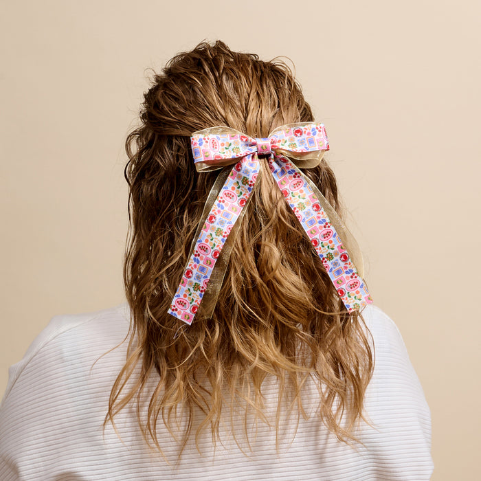 A person with wavy brown hair, seen from behind in a white top, wears The Darling Effect’s Ciao Bella Lovely Layered Hair Bow—an adorable beige accessory with floral and heart patterns.