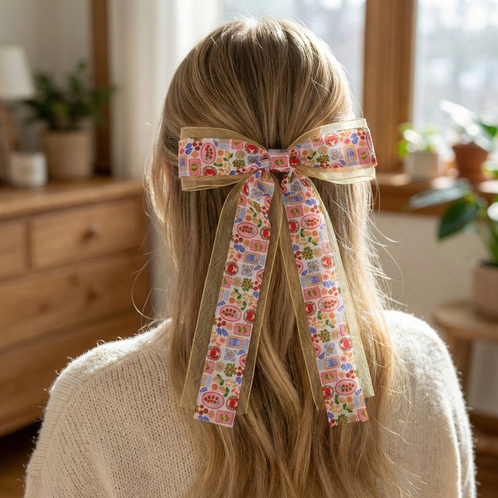A person with long blonde hair wears The Darling Effect’s Ciao Bella Lovely Layered Hair Bow, featuring vibrant floral and geometric patterns, on the back of their head indoors near a wooden dresser and potted plants.