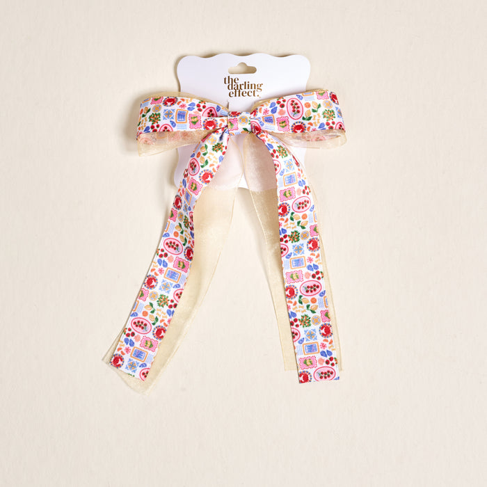 The Ciao Bella Lovely Layered Hair Bow by The Darling Effect features vibrant fruit and floral patterns, pomegranates, blue tiles, gold-trimmed ribbon tails, and a cream backing for a charming accessory.