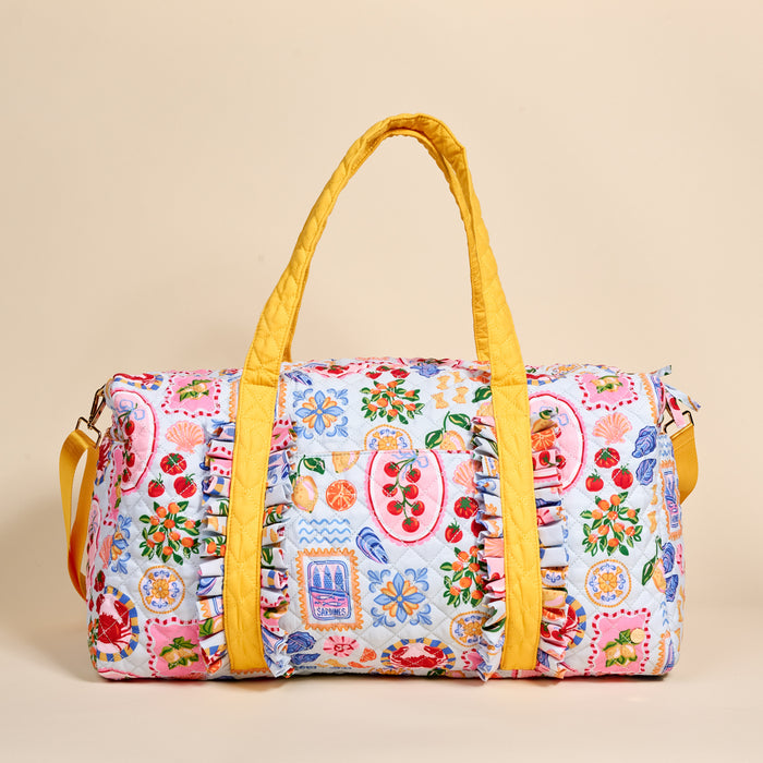 The Darling Effect's Ciao Bella On-the-Go Quilted Weekender Bag features yellow handles, ruffle trim, an adjustable strap, and a vibrant fruit, flower, and sardine print on a light blue background with Mediterranean-inspired motifs.