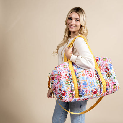 A smiling woman with long blonde hair carries The Darling Effect's Ciao Bella On-the-Go Quilted Weekender Bag, featuring yellow straps, over her shoulder. She wears a light sweater and jeans and stands against a beige background.