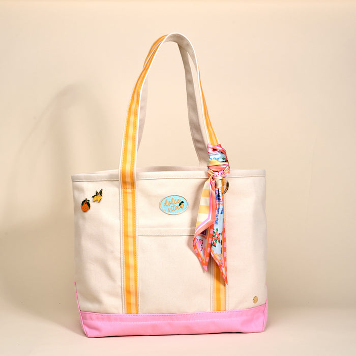 The Darling Effect's Ciao Bella The Darling Canvas Tote Bag features comfy striped handles, a pink base, fruit patches, a round blue Aloha patch, and a colorful scarf on one handle, set on a plain beige background.