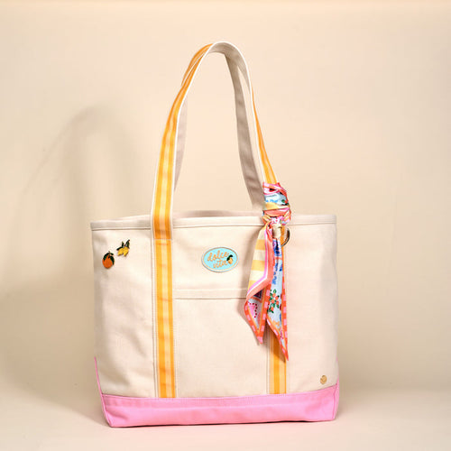 The Darling Effect's Ciao Bella The Darling Canvas Tote Bag features comfy striped handles, a pink base, fruit patches, a round blue Aloha patch, and a colorful scarf on one handle, set on a plain beige background.