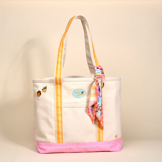 The Darling Effect's Ciao Bella The Darling Canvas Tote Bag features comfy striped handles, a pink base, fruit patches, a round blue Aloha patch, and a colorful scarf on one handle, set on a plain beige background.