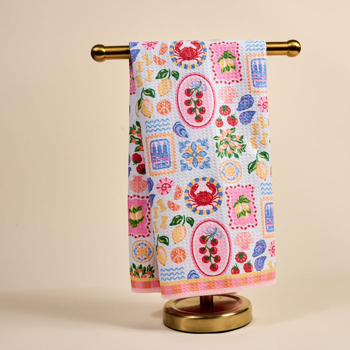 The Darling Effect's Ciao Bella Waffle Tea Towel, adorned with vibrant fruit and seafood prints, hangs neatly on a gold metal stand against a plain beige background.