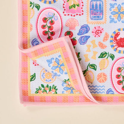 The Darling Effect’s Ciao Bella Waffle Tea Towel features pastel waffle fabric with a pink striped border, adorned with cherries, flowers, shells, lemons, crabs, and sardine cans—an absorbent kitchen essential.