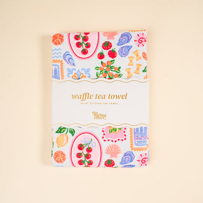 A folded Ciao Bella Waffle Tea Towel by The Darling Effect, featuring colorful fruit and floral patterns and a label, sits on a beige background. This absorbent 18x28" kitchen towel adds charm to any kitchen.