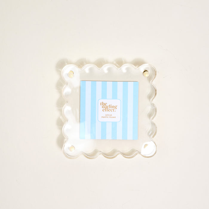 The Darling Effect's Clear Little Moments Acrylic Picture Frame, featuring a square shape, scalloped edges, and four corner holes, displays a blue-and-white striped "the clearing effect" card on a light-colored surface.