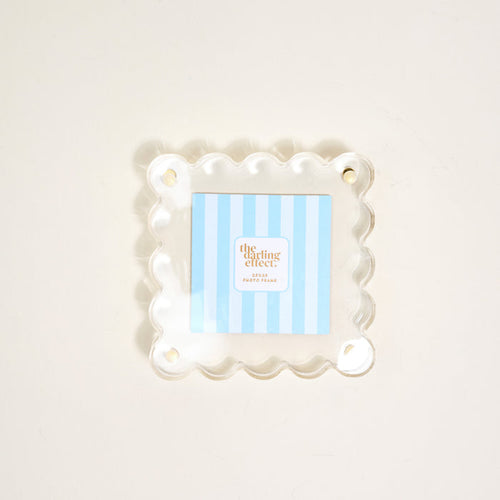 The Darling Effect's Clear Little Moments Acrylic Picture Frame, featuring a square shape, scalloped edges, and four corner holes, displays a blue-and-white striped "the clearing effect" card on a light-colored surface.