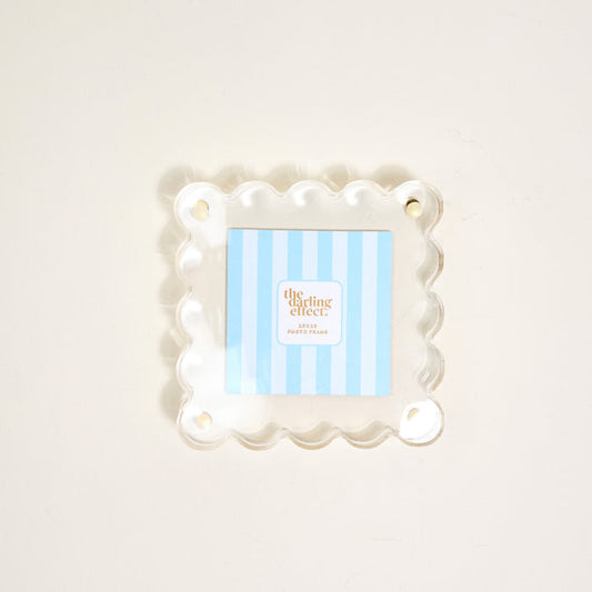 The Darling Effect's Clear Little Moments Acrylic Picture Frame, featuring a square shape, scalloped edges, and four corner holes, displays a blue-and-white striped "the clearing effect" card on a light-colored surface.