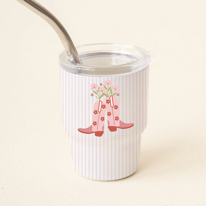 The Darling Effect’s Cowgirl Boots 2 oz Tiny Tumbler features pink vertical stripes and a design of pink rain boots with flowers. It includes a clear lid and reusable metal straw, making it ideal for drinks on the go.