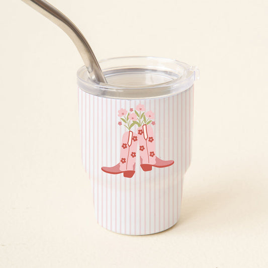 The Darling Effect’s Cowgirl Boots 2 oz Tiny Tumbler features pink vertical stripes and a design of pink rain boots with flowers. It includes a clear lid and reusable metal straw, making it ideal for drinks on the go.
