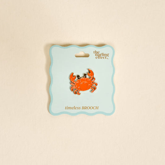 The Darling Effect's Cutie Crab Timeless Brooch, a vibrant orange vintage-inspired enamel pin, is shown on a light blue scalloped card labeled "the darling effect" and "timeless BROOCH" against a beige background.