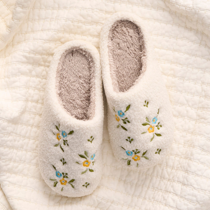 The-darling-effect Dainty Floral Blue & Yellow Fuzzy Slippers, featuring soft interiors and embroidered flowers, rest on a textured cream quilt.
