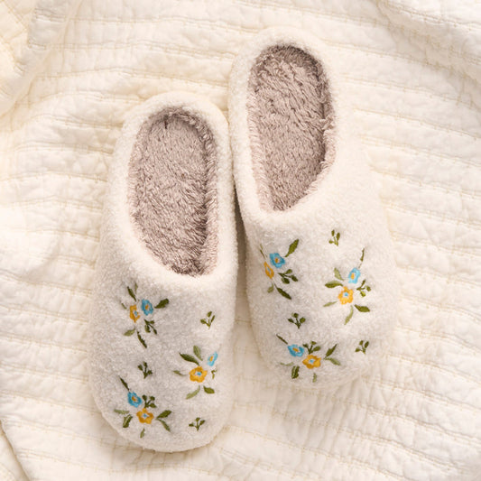 The-darling-effect Dainty Floral Blue & Yellow Fuzzy Slippers, featuring soft interiors and embroidered flowers, rest on a textured cream quilt.