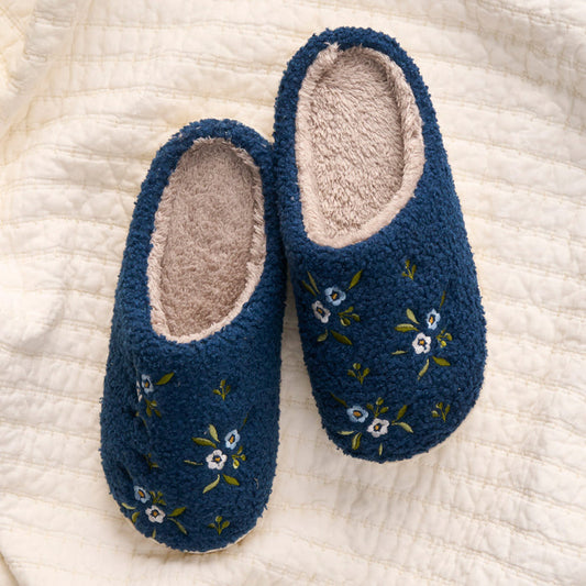 The-darling-effect Dainty Floral Navy Fuzzy Slippers feature white and green embroidered florals, and are shown on a cream quilted surface.